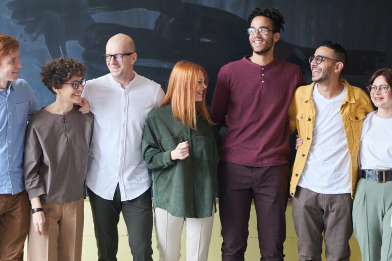 A group of people standing in a row with their arms around each other, smiling and talking to one another.