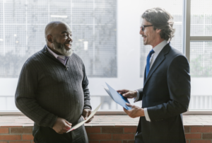 Two men holding papers have a discussion in front of a window.