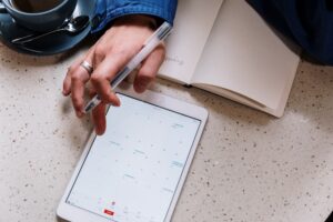 A hand holding a pen hovers over a tablet and an open notebook on a table.