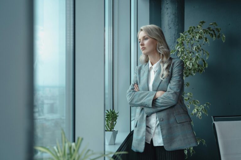 A businesswoman looking through a window in an office, appearing troubled.